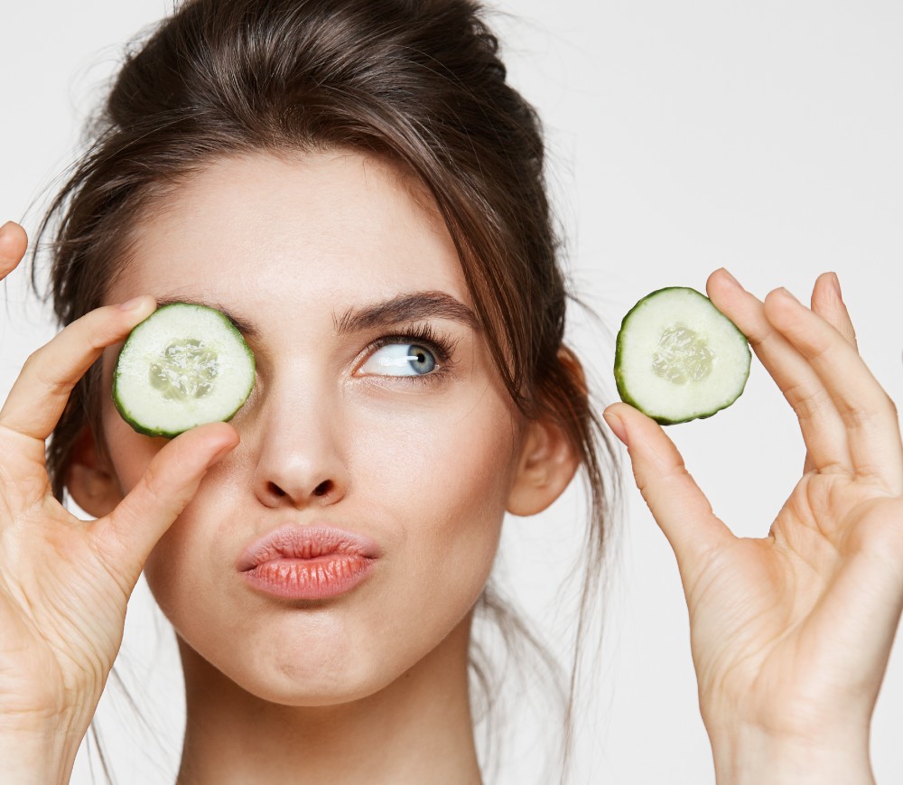 Woman holding cucumber slices near her eyes.